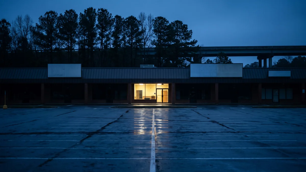 Wide low retail strip beside a highway outlet mall at blue hour in the American South, a modest independent storefront with warm interior light spilling onto wet asphalt, empty parking bays in the foreground, pine treeline behind
