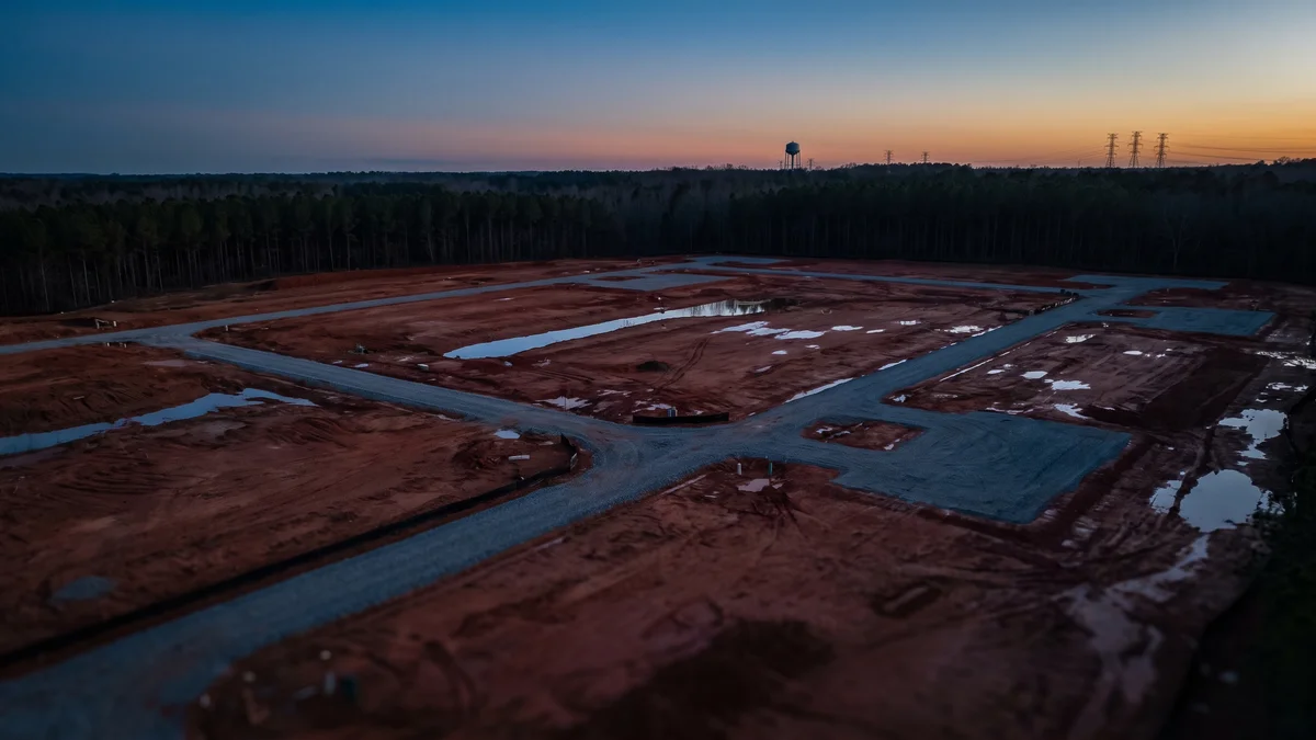 Wide aerial-style view of cleared red-clay industrial land at the edge of dense Piedmont pine forest at dusk, graded pads and access roads cut into the earth, a distant water tower and power lines on the horizon, deep blue sky